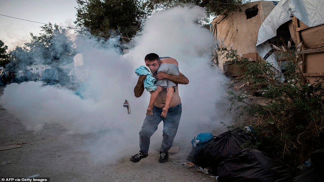 رجل يحمل فتى أثناء اشتباكات مع الشرطة خارج مخيم للاجئين في ليسبوس اليونانية، وأطلقت الشرطة الغاز المسيل للدموع للسيطرة على الحشود التي بدأت أعمال الشغب بعد اندلاع الحريق داخل المخيم المكتظ